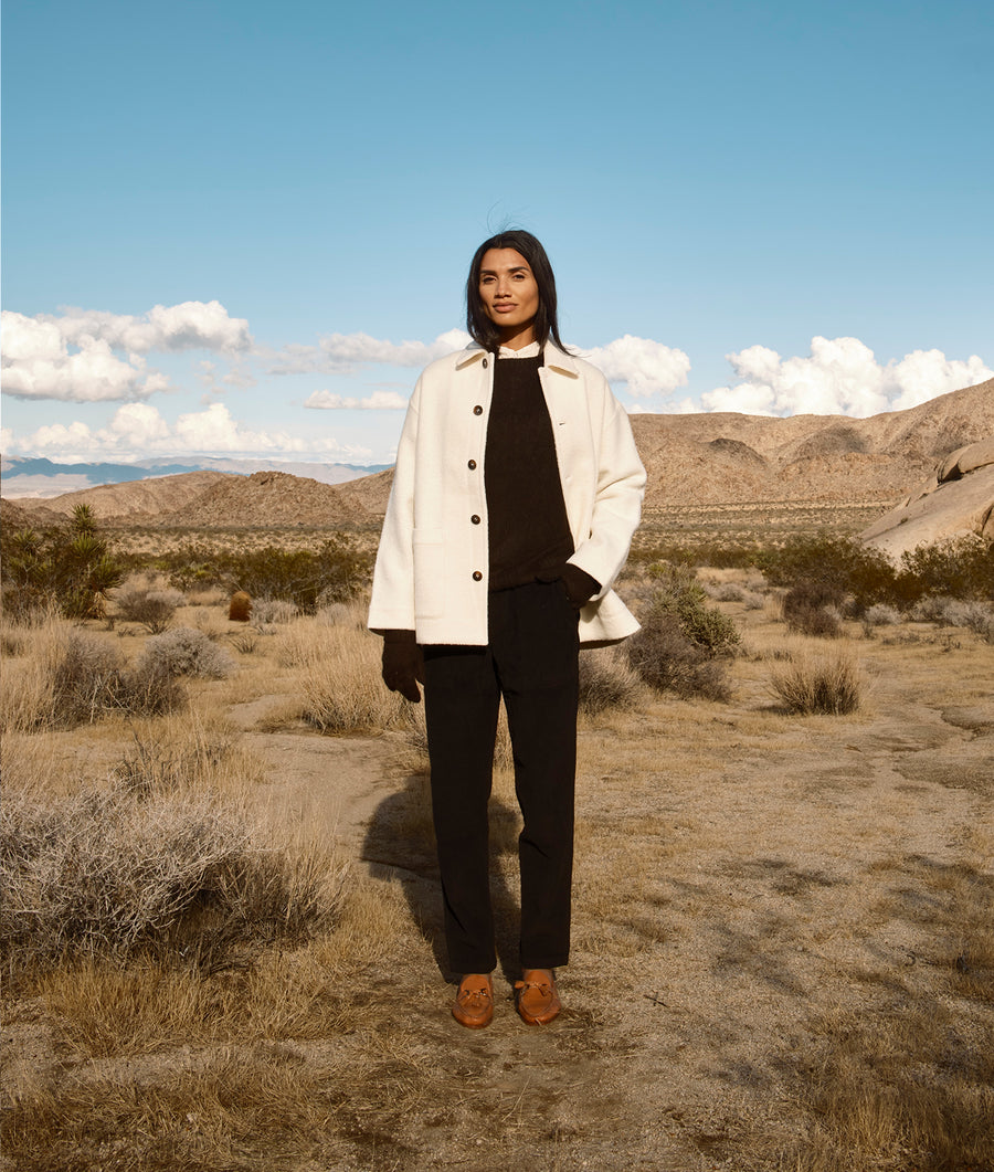 A model wearing a white button-up coat over a black outfit, standing in a desert landscape with mountains in the background - Industry of All Nations