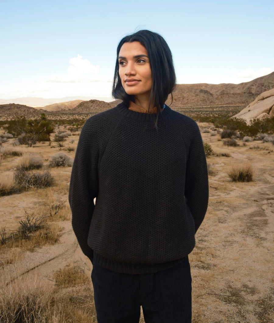A woman wearing a black knit sweater in a desert landscape with mountains in the background - Industry of All Nations