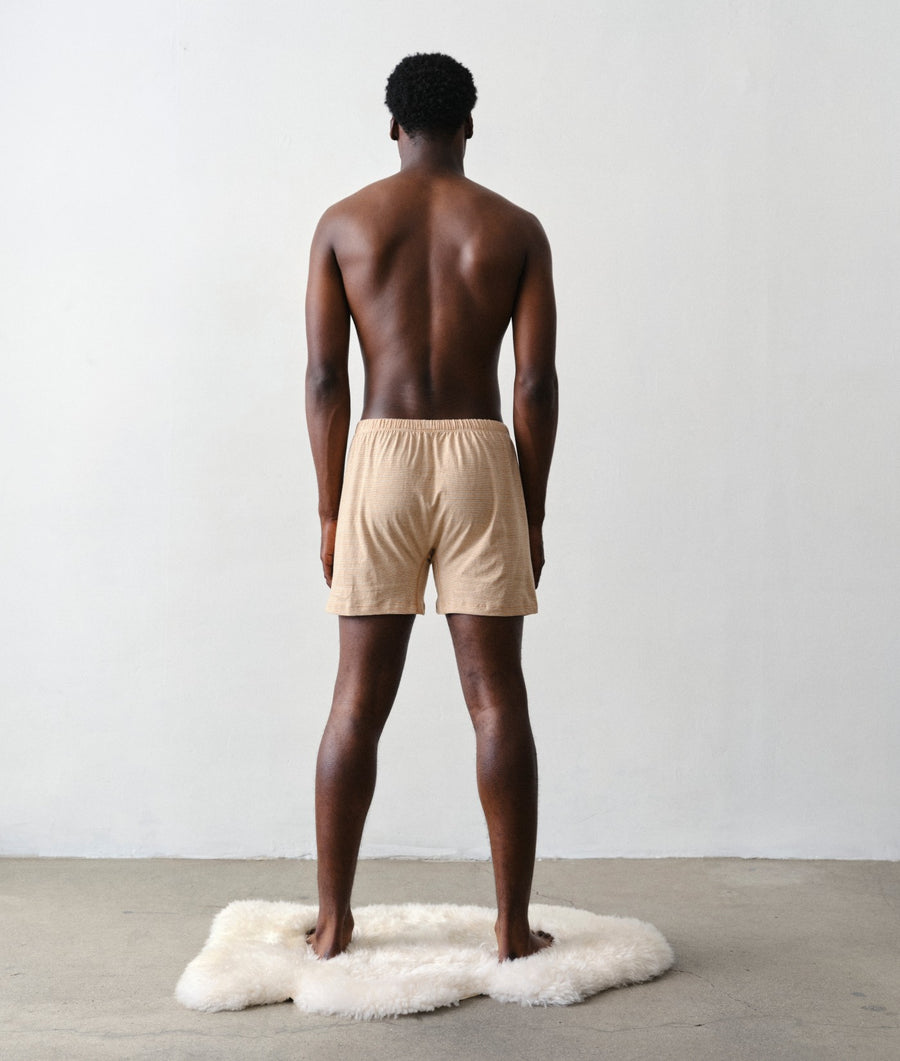 A shirtless man stands with his back to the camera on a white sheepskin rug, wearing IOAN Wild Boxers in a minimalist studio with a white wall and concrete floor.