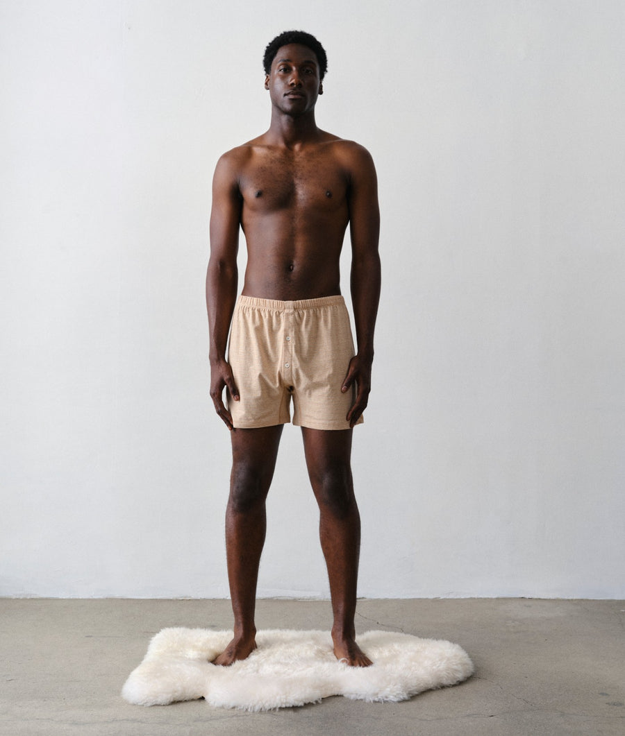 A shirtless man stands facing forward on a white sheepskin rug, wearing IOAN Wild Boxers in a minimalist studio with a white wall and concrete floor.