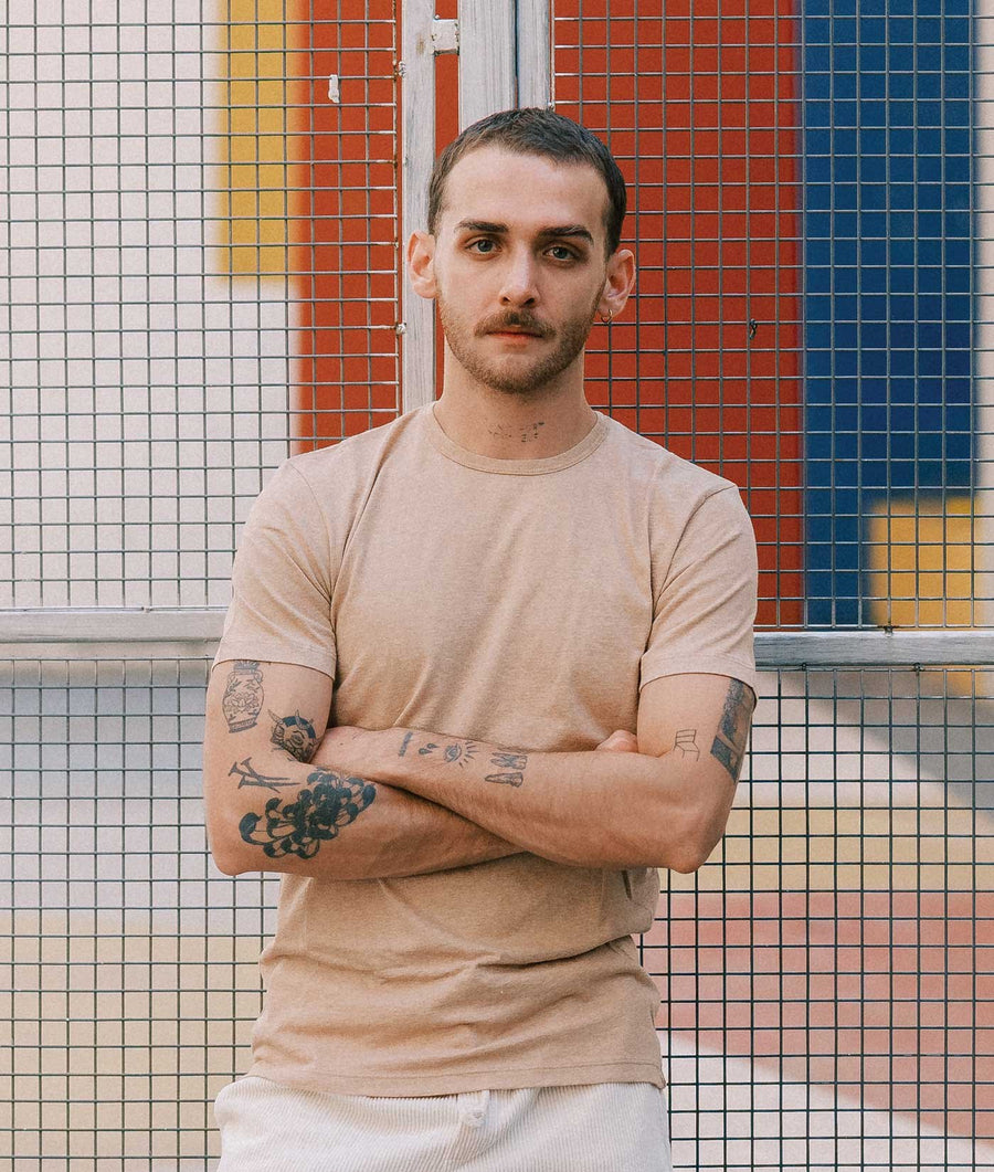A tattooed man stands with his arms crossed in front of a wire grid fence, wearing an IOAN Wild T-Shirt in Caramel with white pants, against a background of red, blue, yellow, and white panels.
