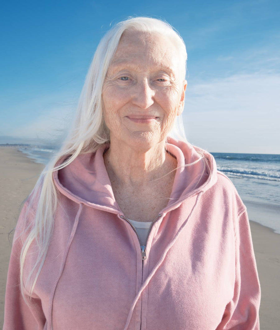 The image shows an elderly woman with long white hair wearing a pink zip-up hoodie, standing on a beach with a blue sky in the background. - Industry of All Nations