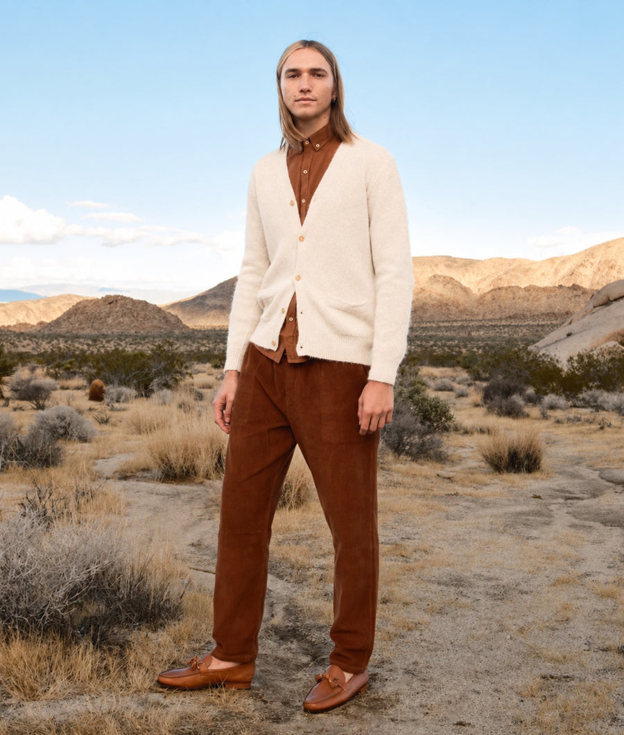 A man stands in a desert landscape wearing a cream knit cardigan over a brown button-up shirt, brown corduroy trousers, and brown loafers.