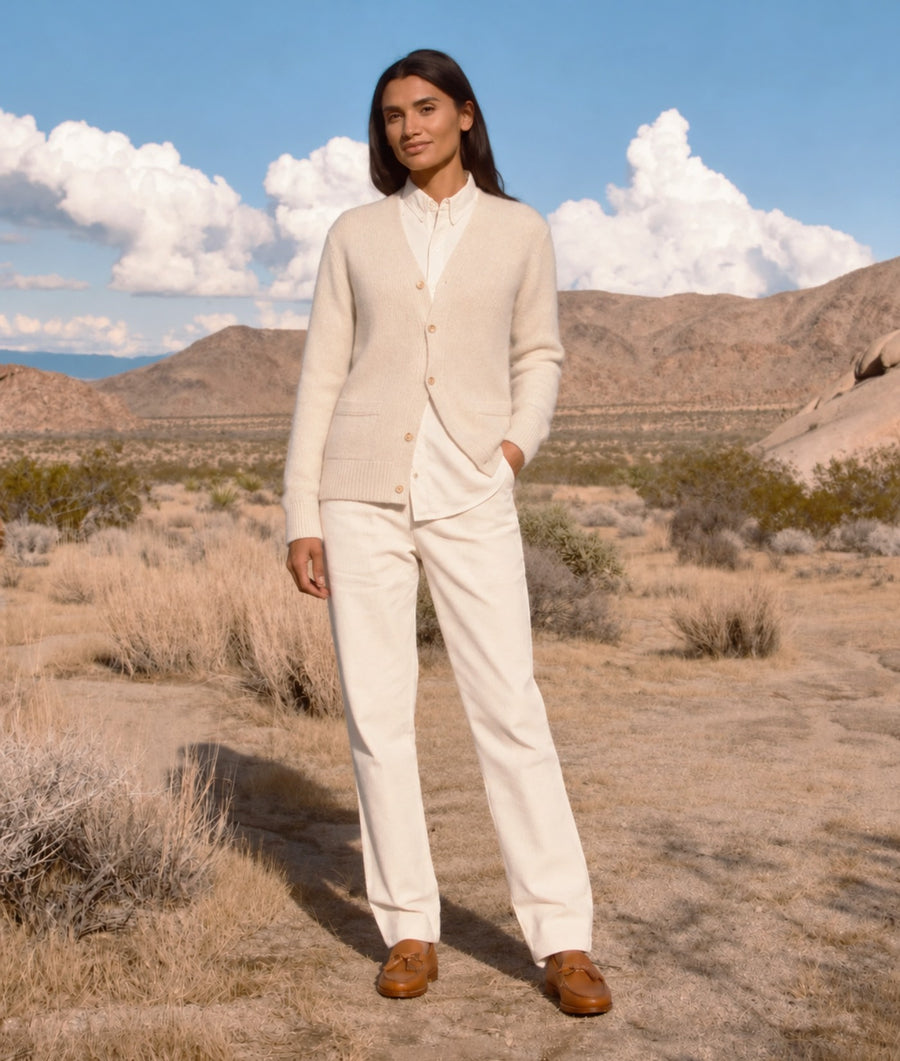 A woman stands in a desert landscape wearing a cream-colored knit cardigan over a white collared shirt, matching cream trousers, and brown suede loafers.