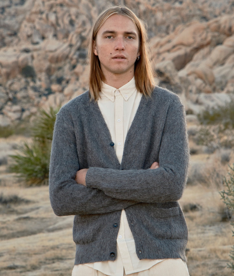 A man with long light brown hair stands in a rocky desert landscape wearing a gray button-front cardigan over a cream button-up shirt.