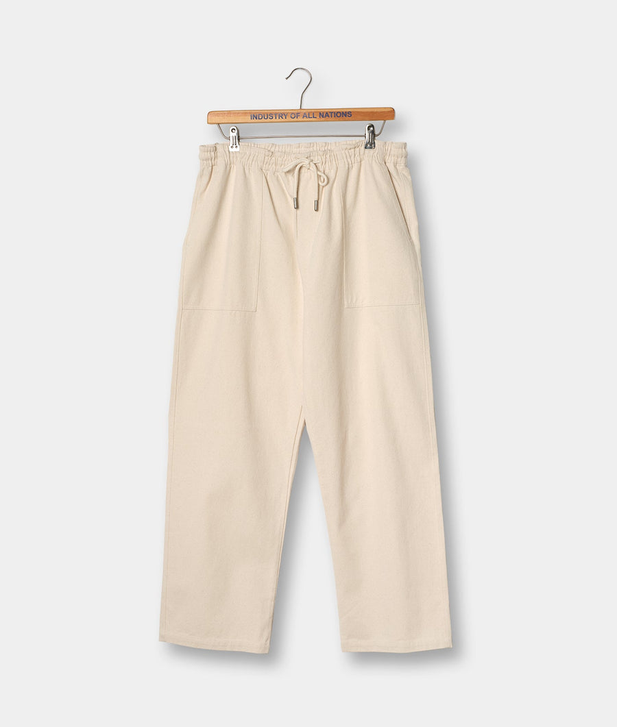 Cream-colored relaxed fit pants with drawstring waist, displayed hanging on a wooden hanger with "Industry of All Nations" text - Industry of All Nations
