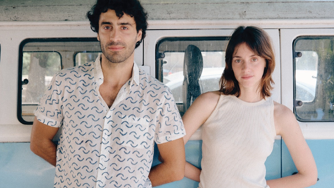 A man and a woman standing side-by-side in front of a vintage-style blue and white bus. The man on the left wears a white short-sleeve button-up shirt with a blue wavy pattern, and the woman on the right wears a cream-colored ribbed sleeveless top.