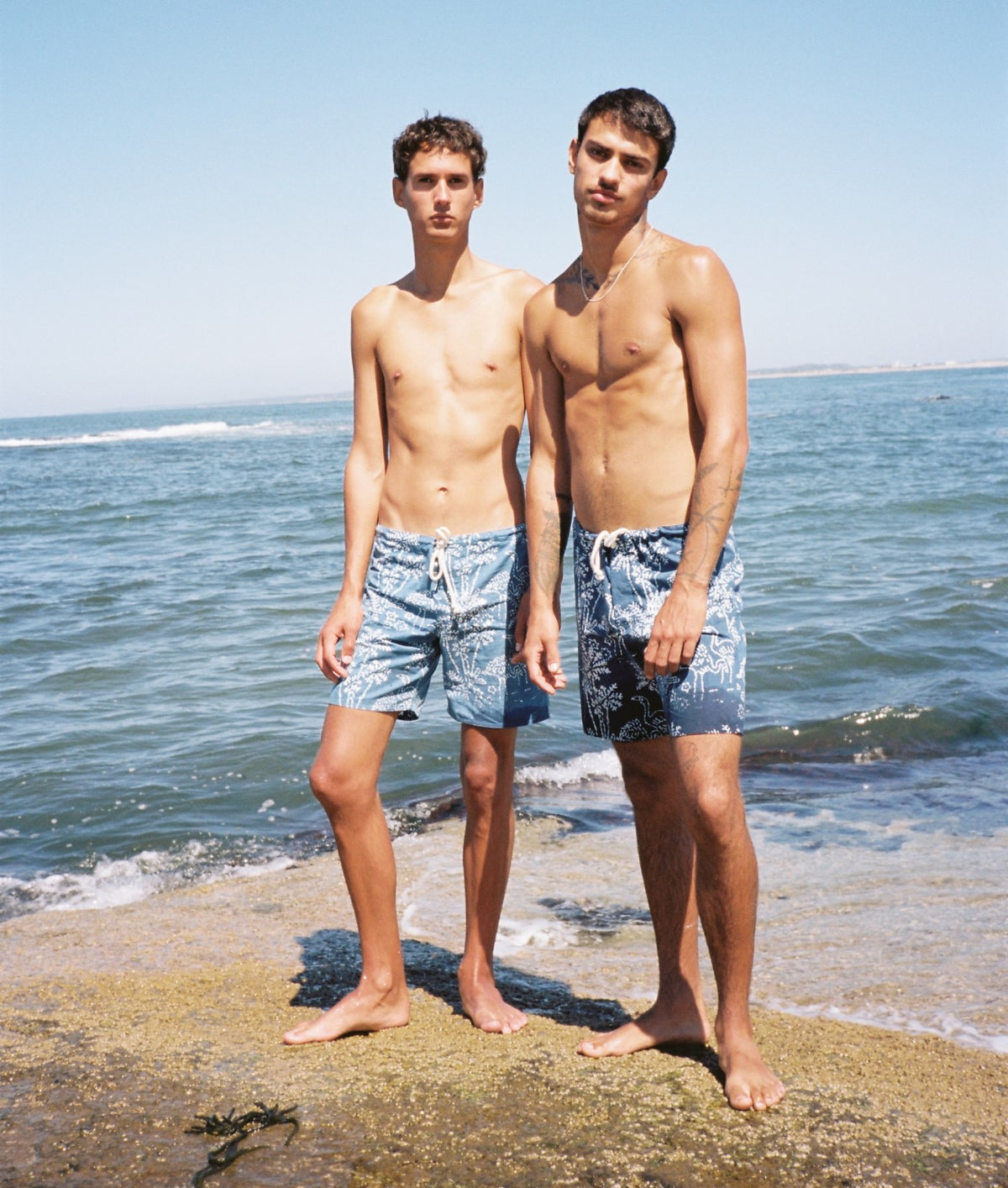 Two shirtless men standing on a sandy beach wearing blue swim trunks, with the ocean and blue sky in the background.