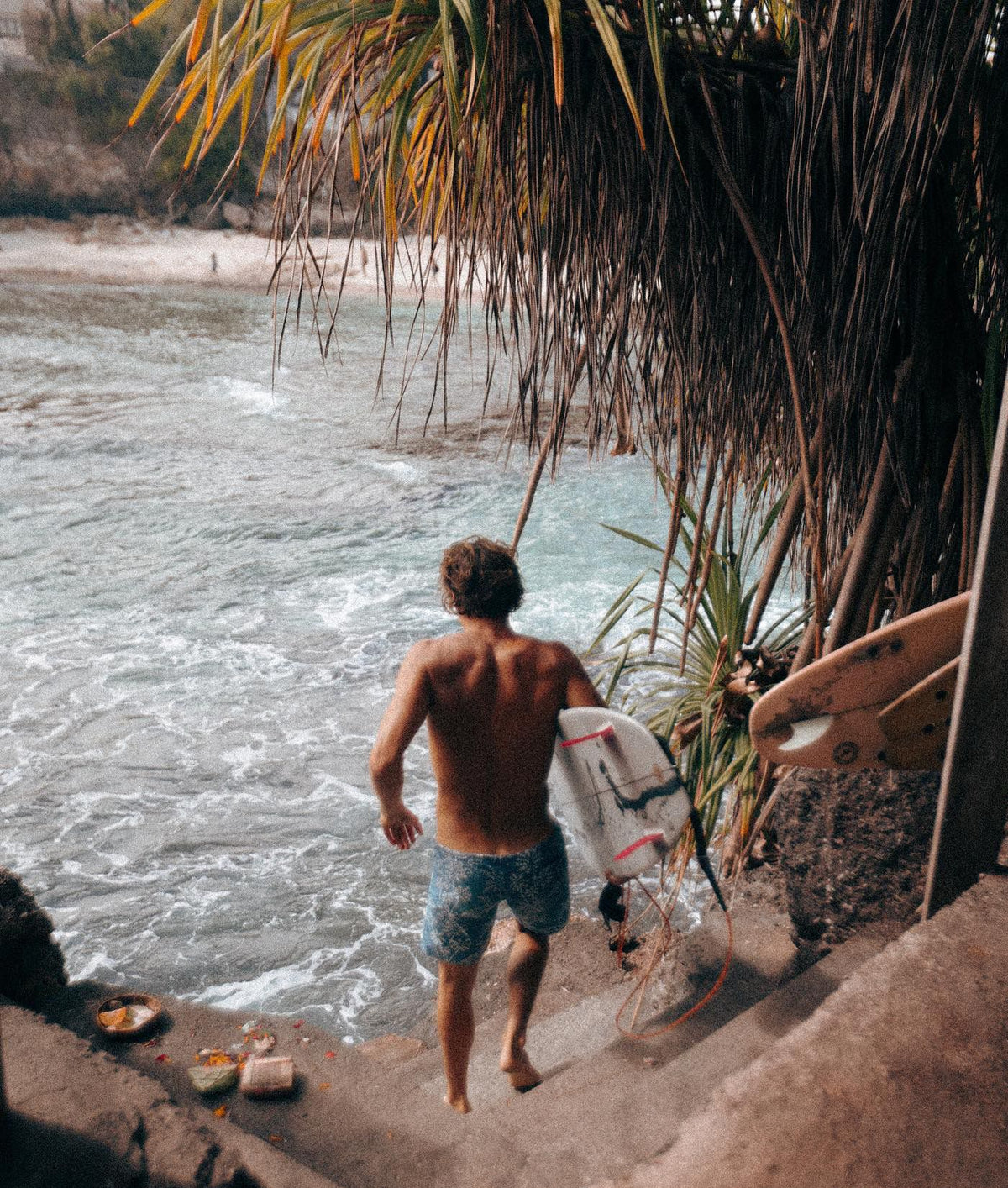 A shirtless man carrying a white surfboard down stone steps toward the ocean, framed by palm leaves and rocks, with a second surfboard visible on the right.