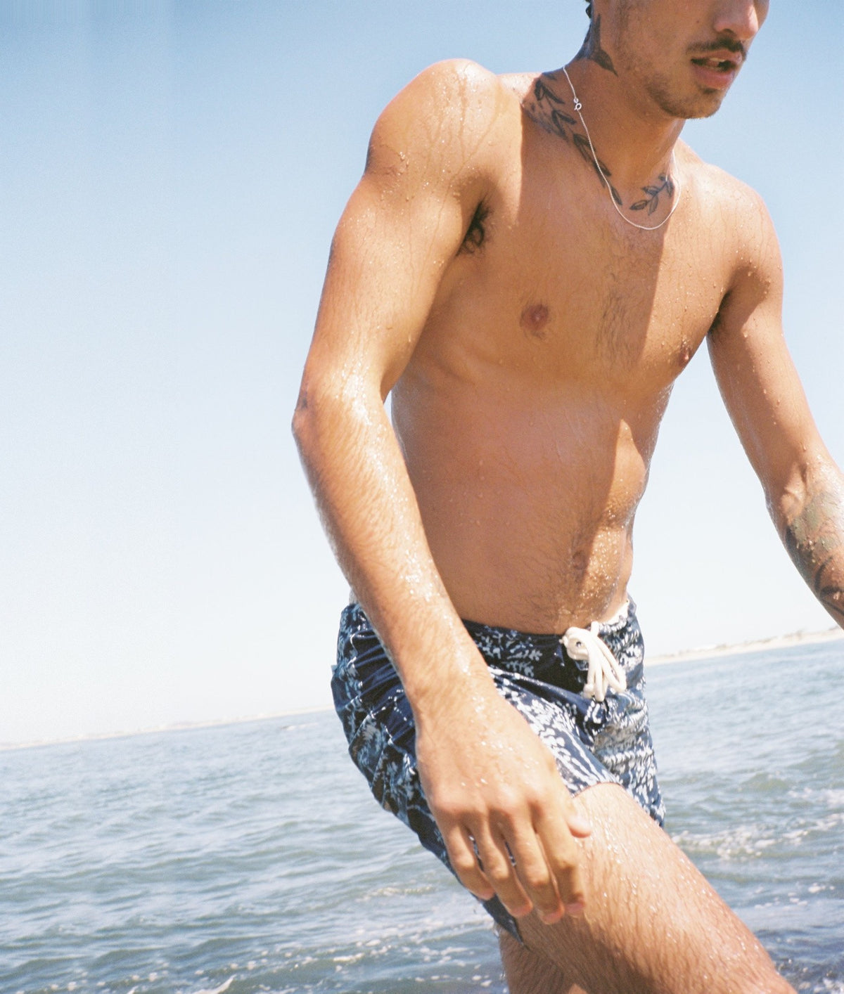 A man standing in shallow water wearing dark swim trunks with a light pattern, photographed outdoors under a clear blue sky.