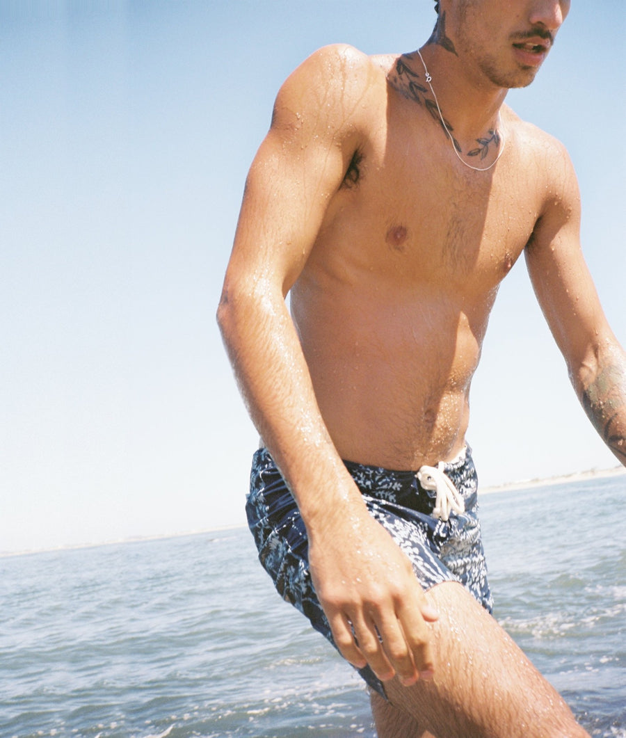 A man standing in shallow water wearing dark swim trunks with a light pattern, photographed outdoors under a clear blue sky.