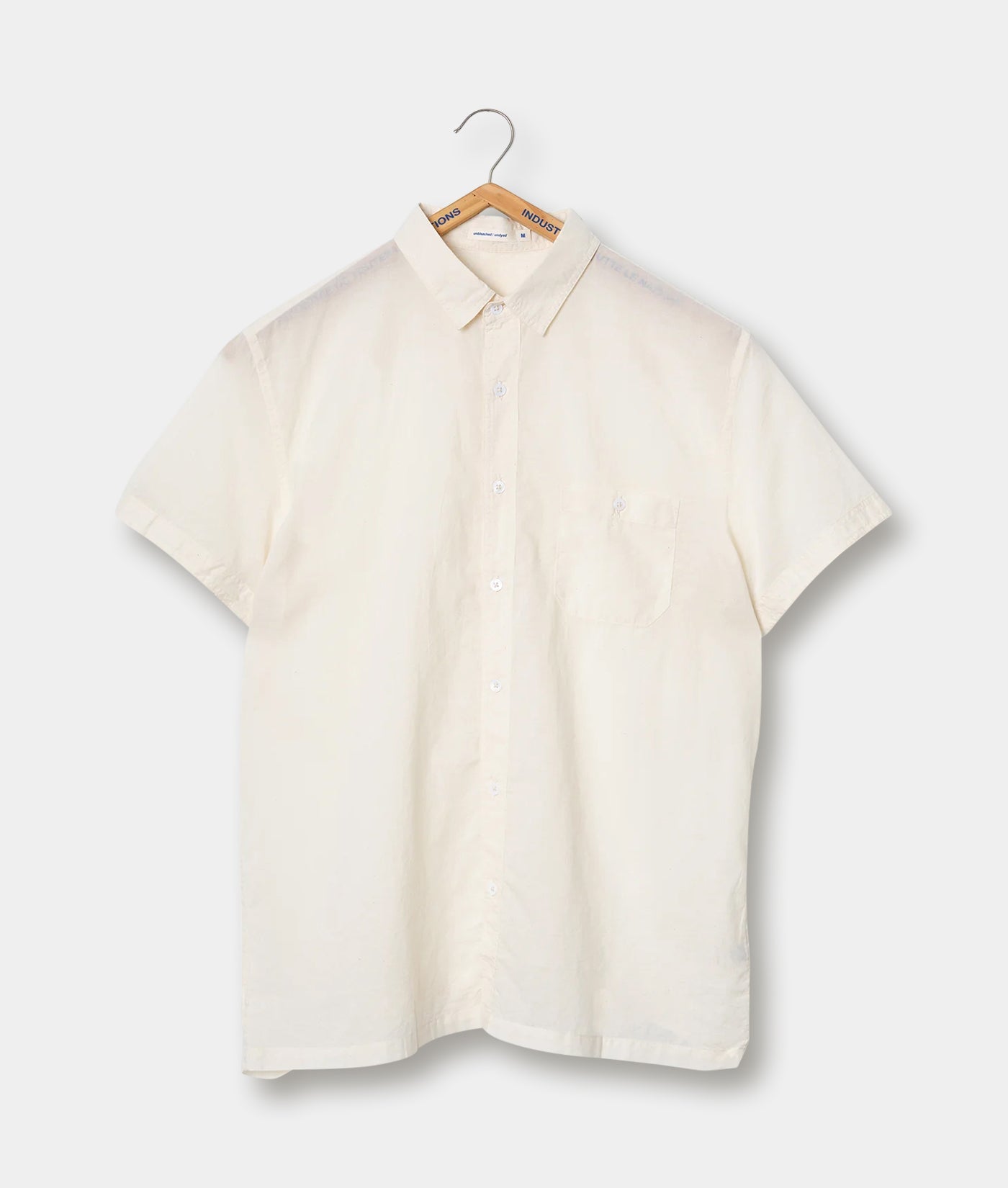 Cream-colored button-down shirt, hung on a wooden hanger against a plain background - Industry of All Nations