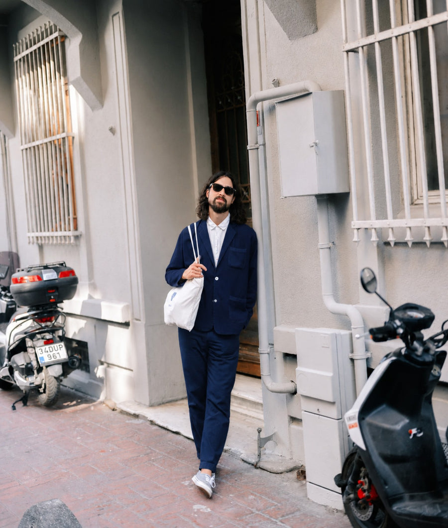 A man wearing a navy blue two-piece suit, white shirt, and sunglasses walks down a street next to a building, carrying a white shoulder bag.