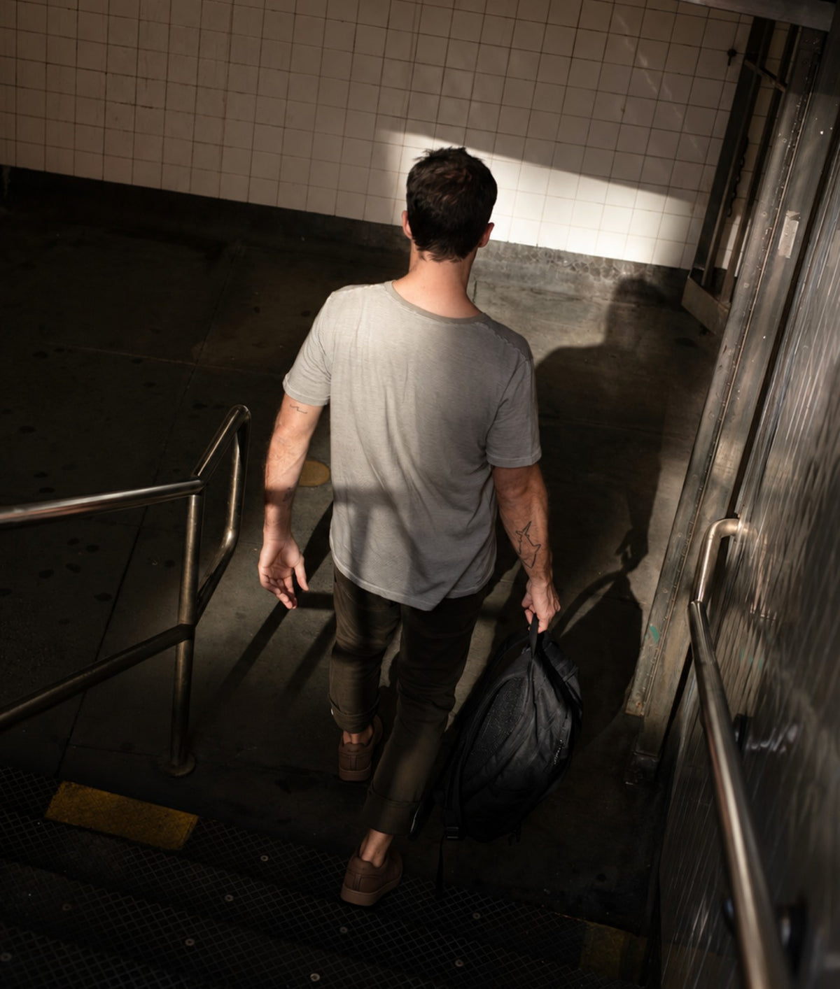 A man seen from behind walking down tiled stairs in a subway or station, wearing a light gray t-shirt, dark pants, and brown shoes while carrying a black duffel bag.