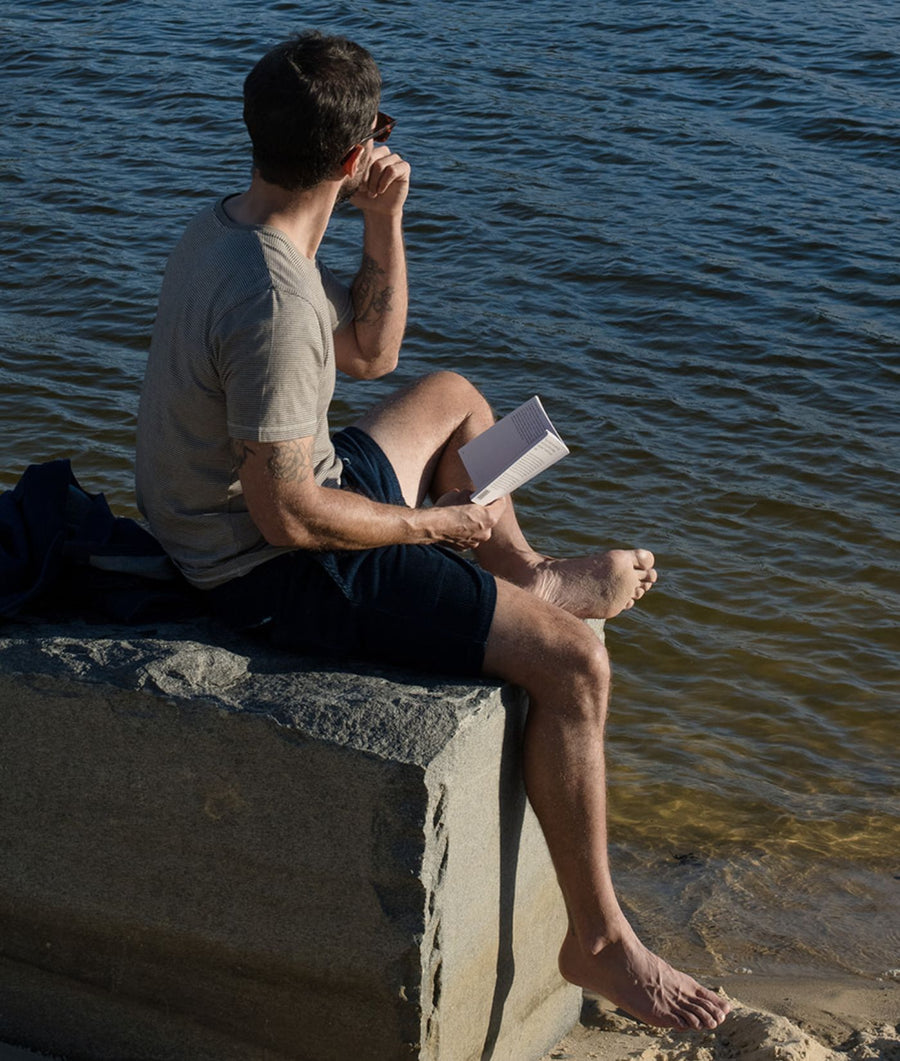 A man sits barefoot on a concrete pier by the water, wearing a light beige t-shirt and dark shorts, holding a book and looking out toward the water.
