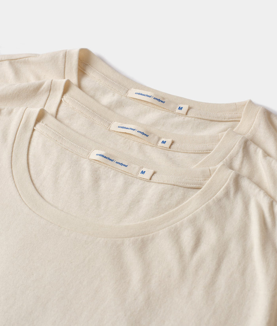 A close-up detail shot of three cream-colored undyed cotton t-shirts with the brand name "unbleached/undyed" printed on the neck tags - Industry of All Nations.