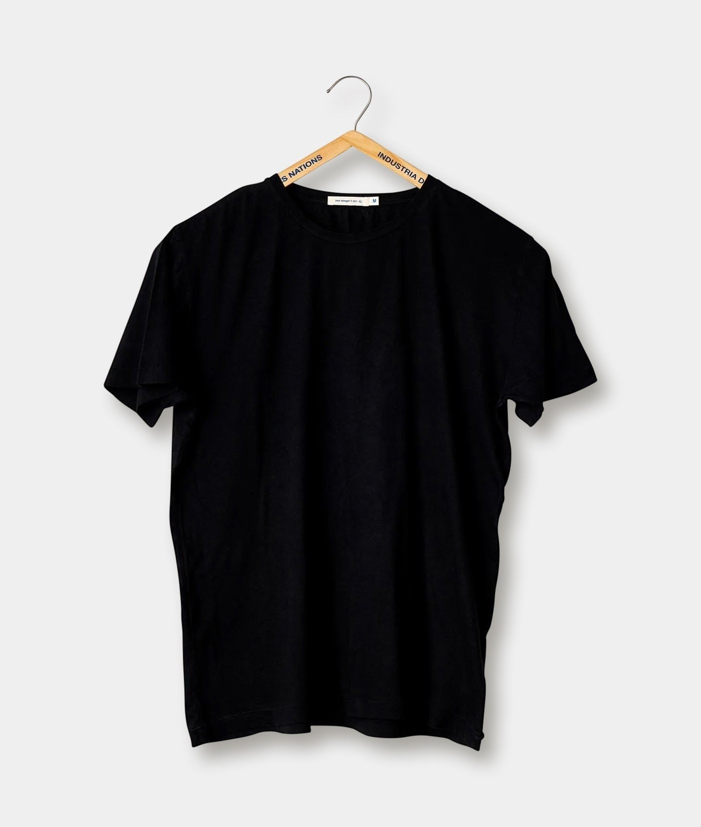 Black t-shirt hanging on a wooden hanger with the brand name "Industry of All Nations" visible - Industry of All Nations