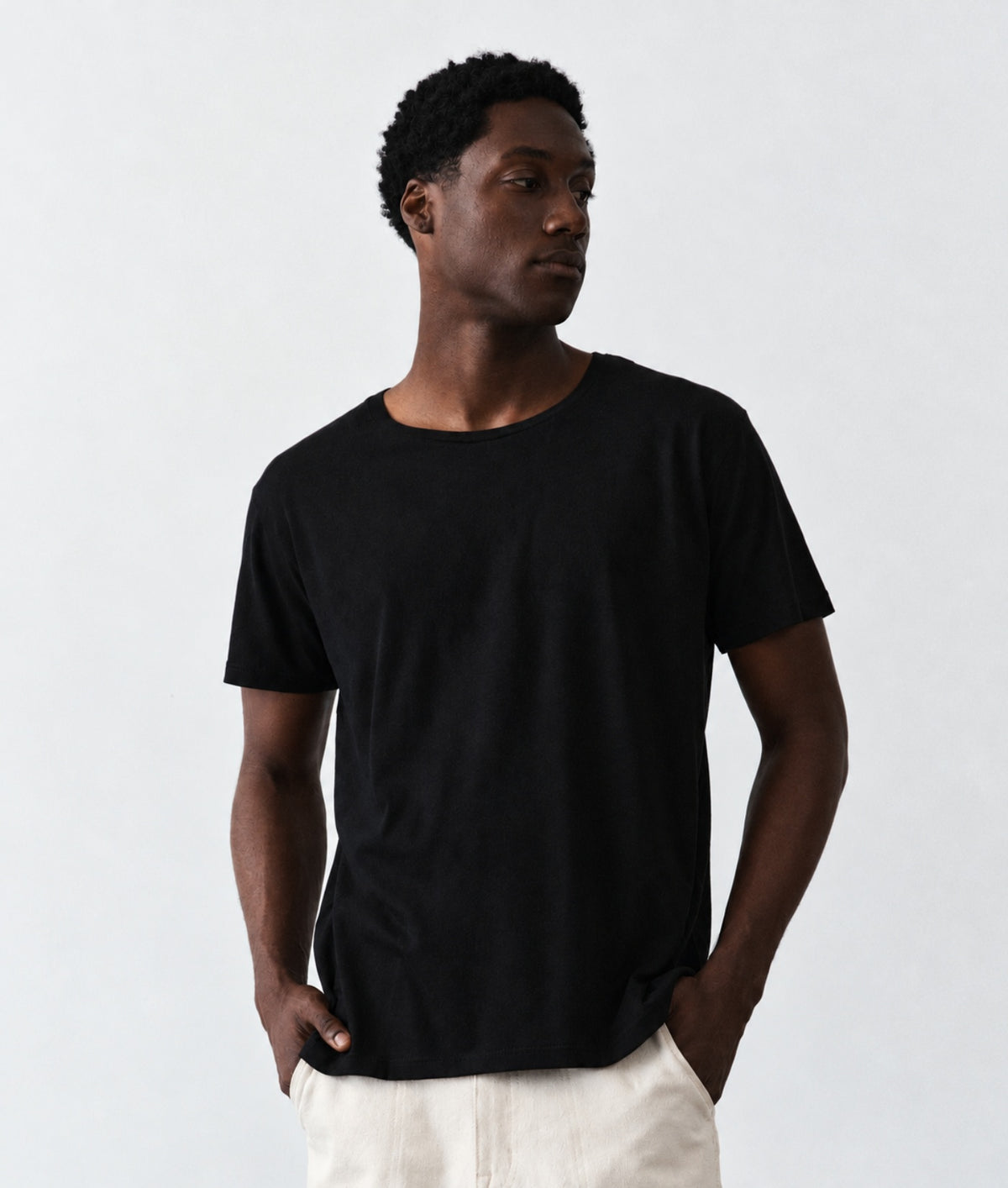 A man wearing a plain black short-sleeve crewneck t-shirt and off-white trousers, standing against a light gray studio background.