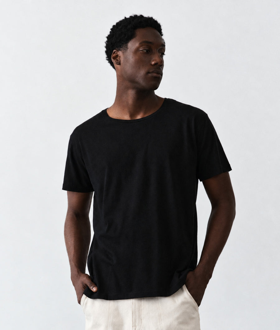 A man wearing a plain black short-sleeve crewneck t-shirt and off-white trousers, standing against a light gray studio background.