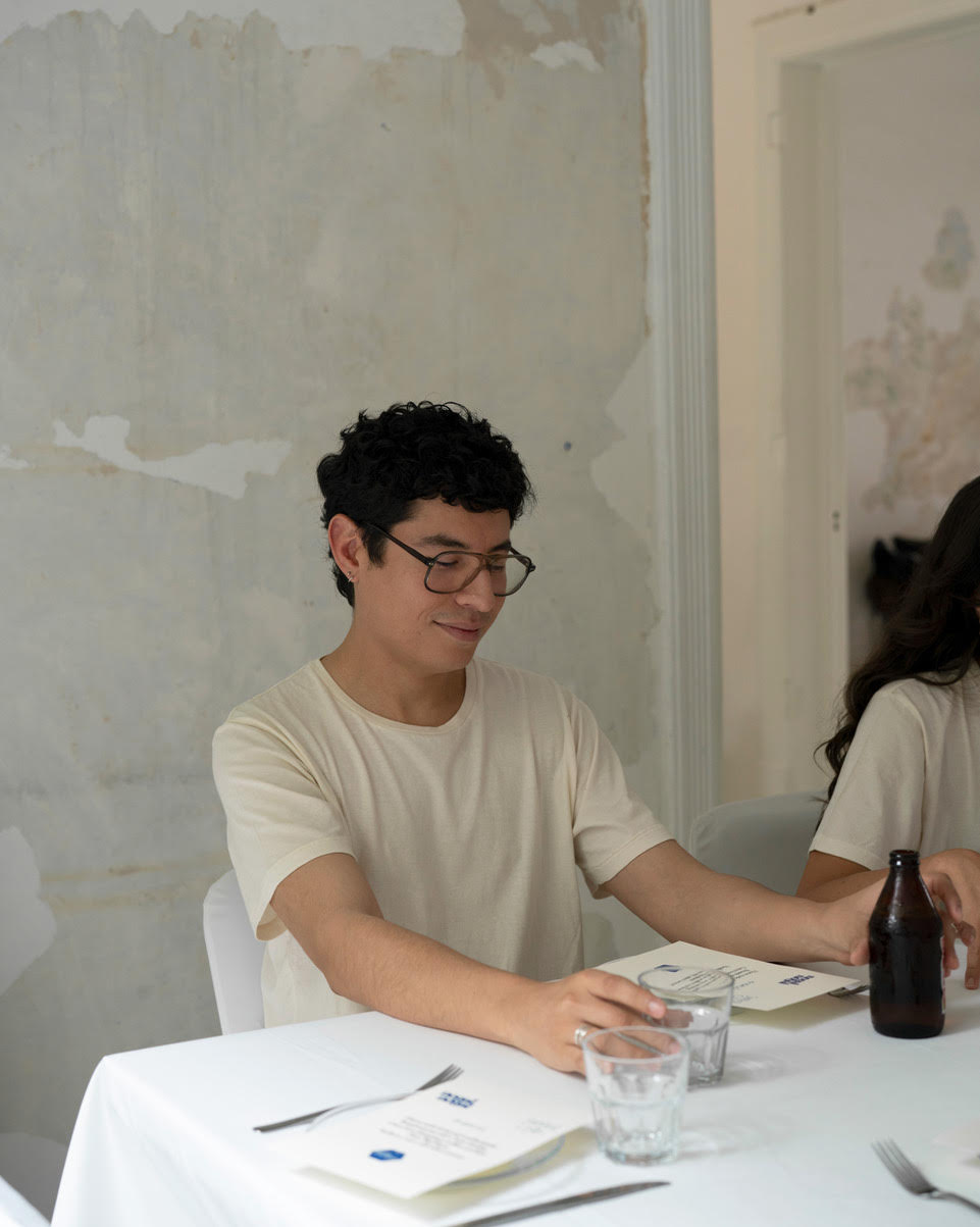 A man wearing IOAN undyed t-shirt and glasses sits at a white table with water glasses, a bottle, and printed materials in a minimalist, modern interior setting.