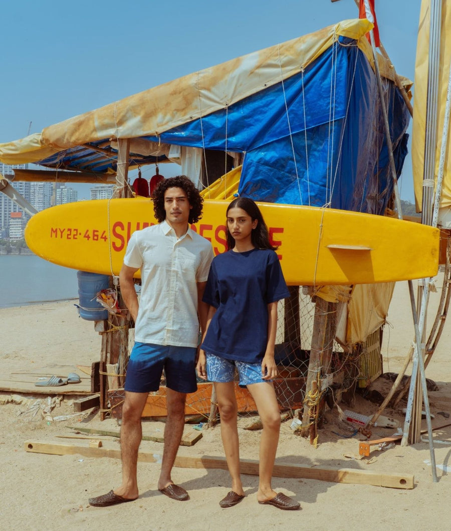 A man and a woman stand on a sandy beach in front of a makeshift beach shack with a large yellow surfboard. The man wears a white short-sleeve button-up shirt and navy shorts, while the woman wears a navy t-shirt and patterned shorts.