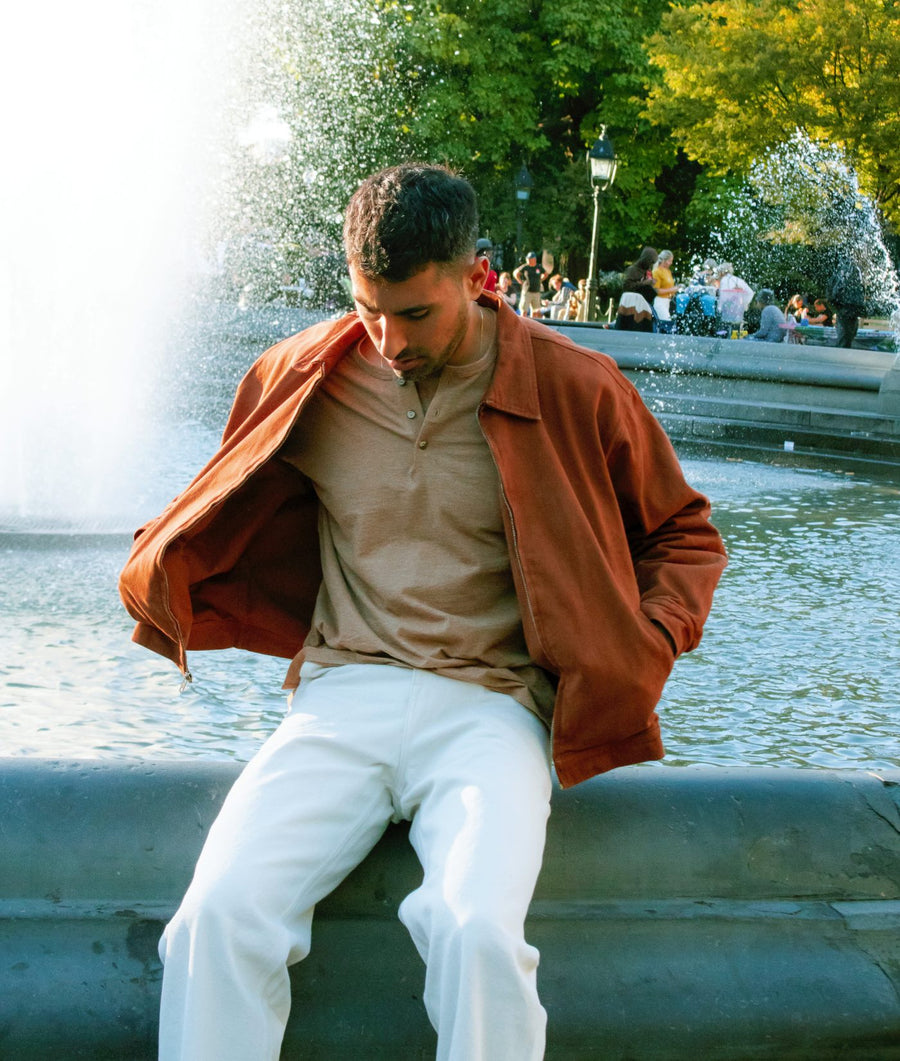 A man sitting on the edge of a fountain in a park, wearing an IOAN Kamaleon Jacket in Acacia over a beige polo shirt and white trousers.