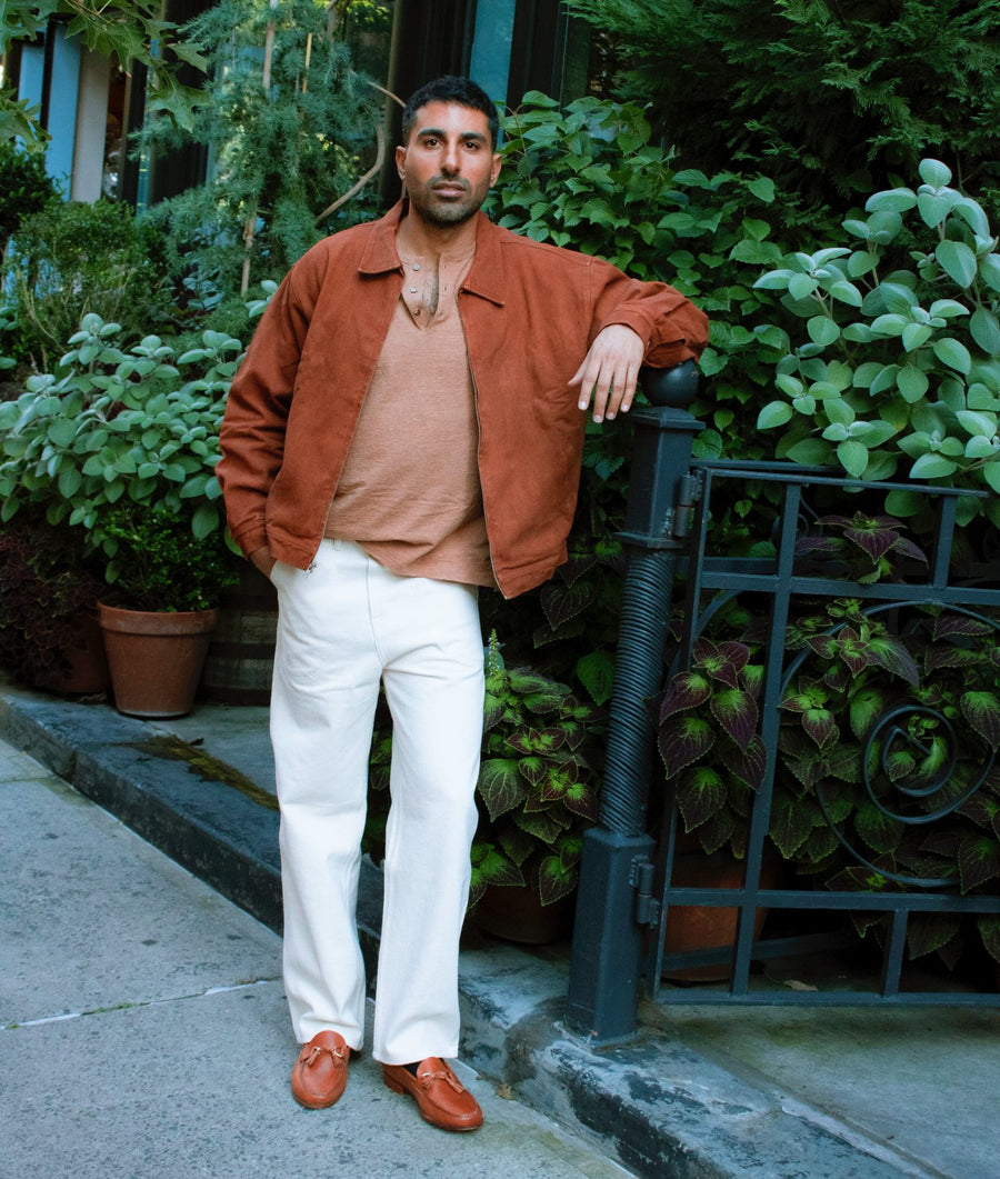 A man stands on a sidewalk, leaning against a black metal fence, wearing an IOAN Kamaleon Jacket in Acacia over a tan knit sweater, white trousers, and brown leather loafers.