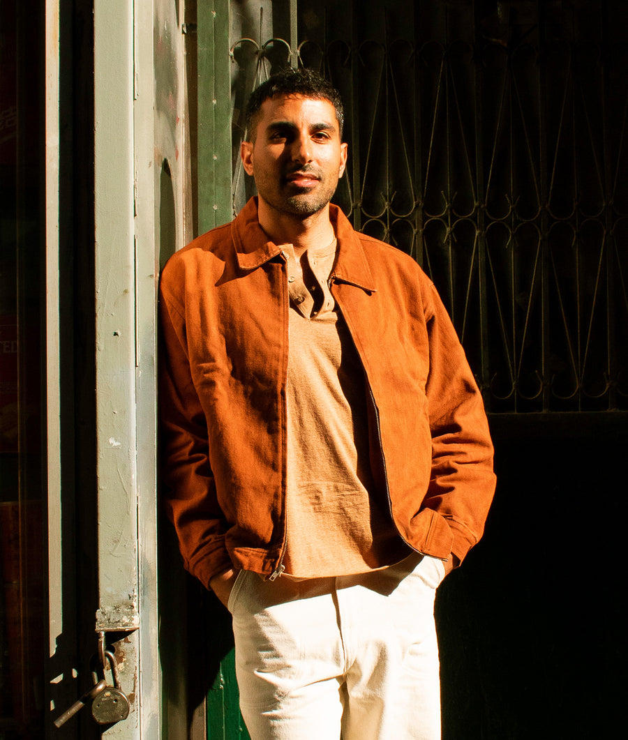 A man in an orange cotton jacket standing in front of a dark green door - Industry of All Nations