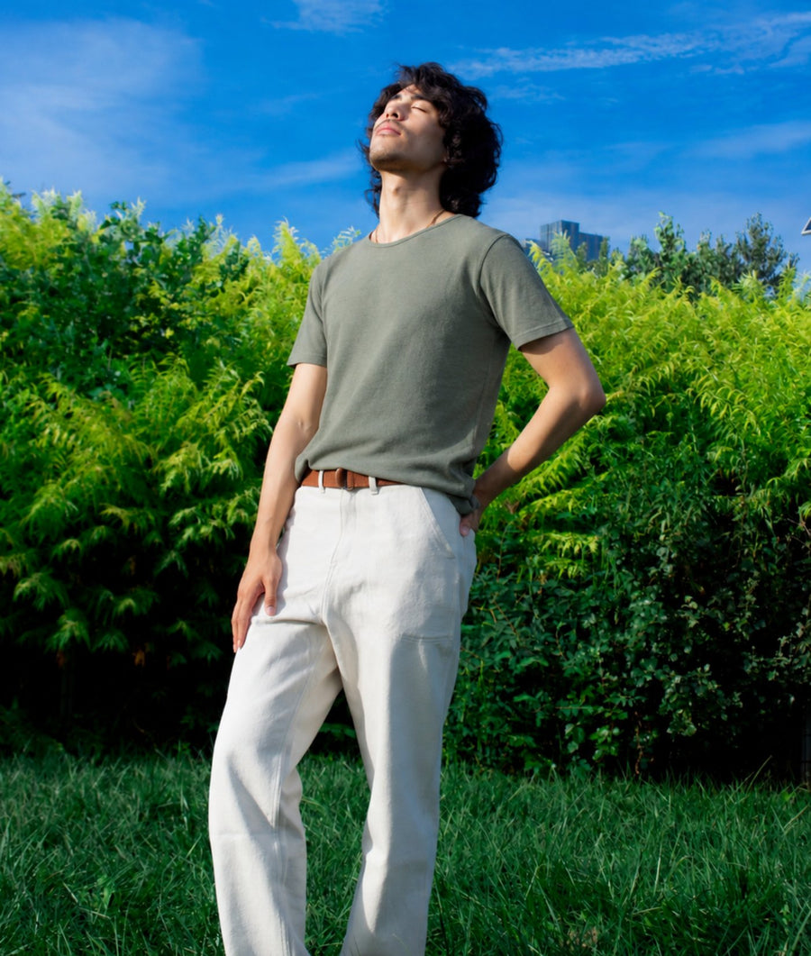 A man wearing an olive green crewneck t-shirt and off-white trousers stands outdoors in a grassy park with a green hedge and blue sky in the background.