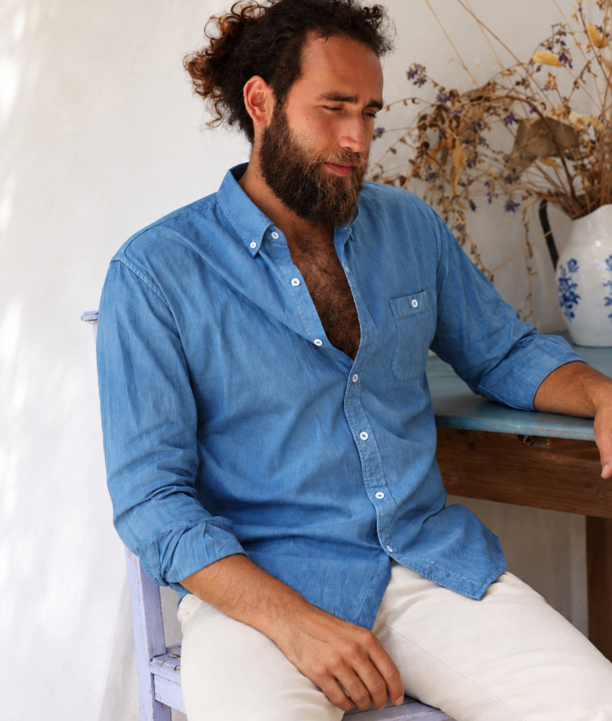A man with a beard and curly hair wearing IOAN Indigo 6 button-down shirt and off-white trousers, sitting in a rustic indoor setting.