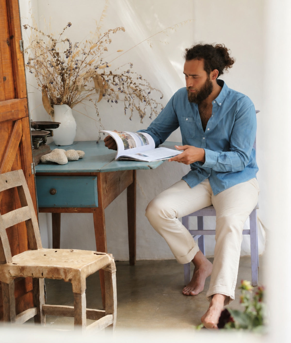 A man with a beard and curly hair sits barefoot on a purple chair, reading an open magazine. He is wearing an IOAN Madras Shirt in Indigo 6 and cream trousers in a rustic, sunlit room with a blue wooden table, a wooden chair, and a white curtain in the background.