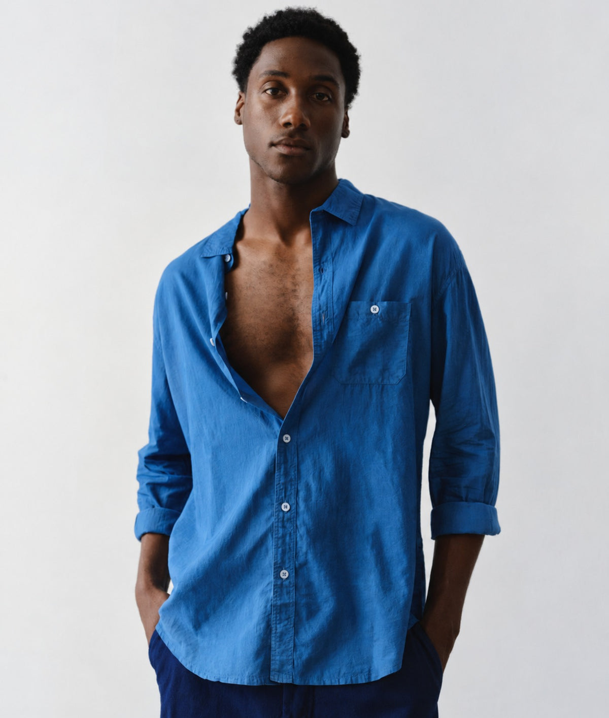 A man wearing a medium blue long-sleeve button-down shirt with a chest pocket, standing against a plain light gray background.