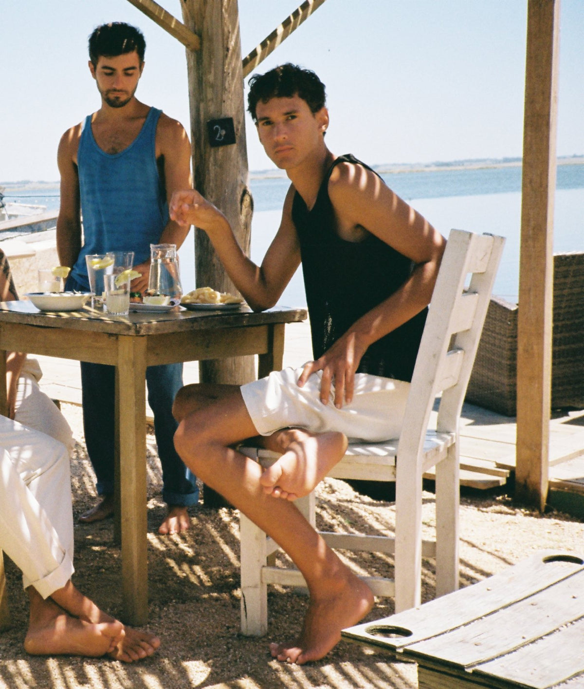 Two men sitting and standing around a wooden table at an outdoor beachside cafe or bar, with water and a wooden structure visible in the background. One seated man wears a black tank top and white shorts, and another man stands wearing a blue tank top and dark pants.