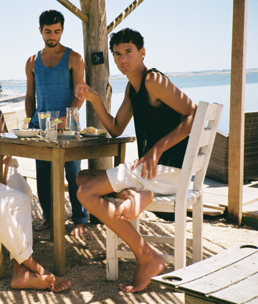 Two men sitting and standing around a wooden table at an outdoor beachside cafe or bar, with water and a wooden structure visible in the background. One seated man wears a black tank top and white shorts, and another man stands wearing a blue tank top and dark pants.