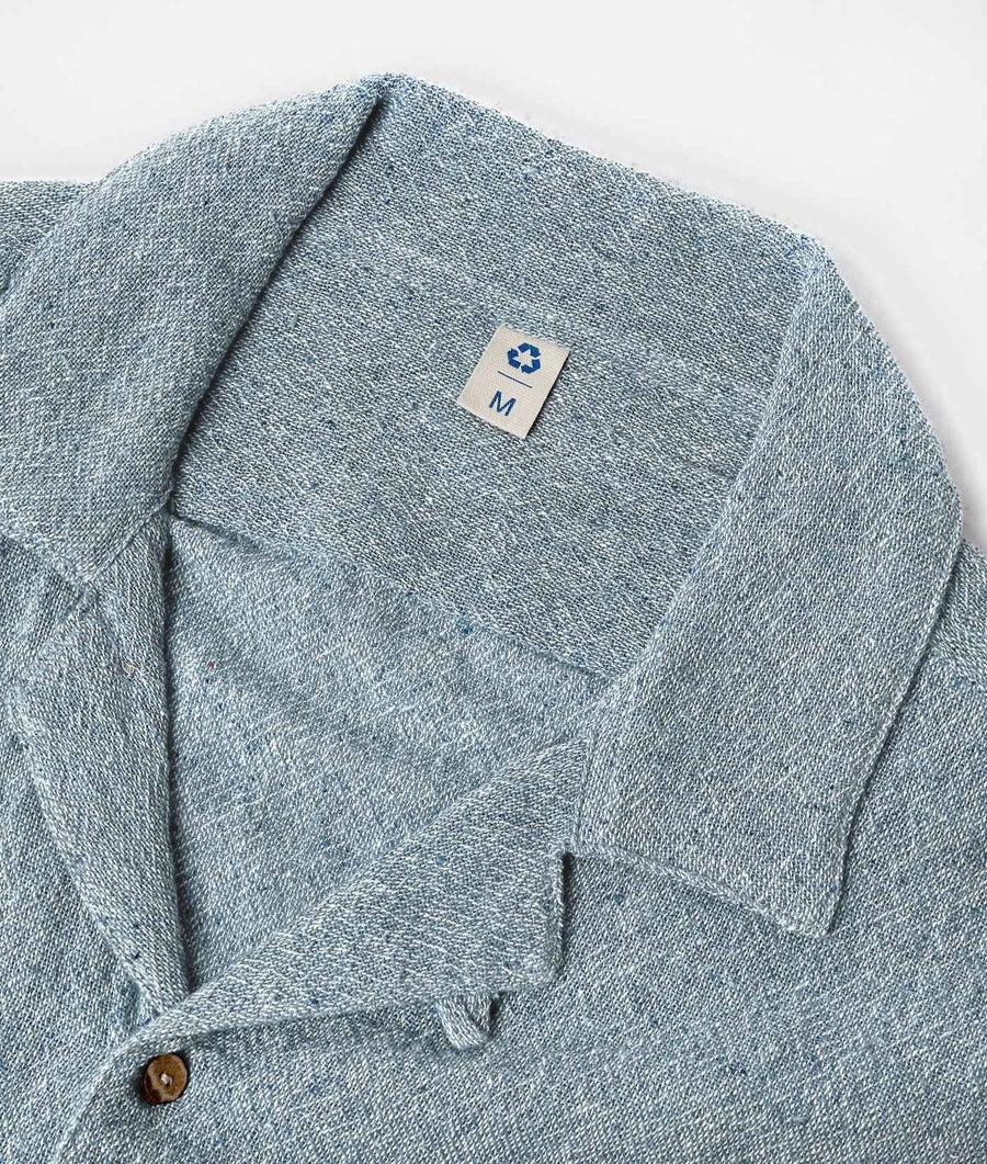 Close-up of a light blue-gray textured knit collared shirt with a small fabric size tag reading M on the inner collar, photographed on a clean white background.