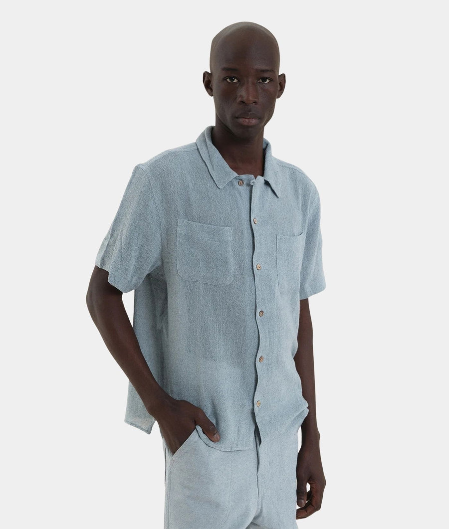 A man wearing a light blue short-sleeve button-up shirt and matching light blue pants, standing against a plain light gray studio background.