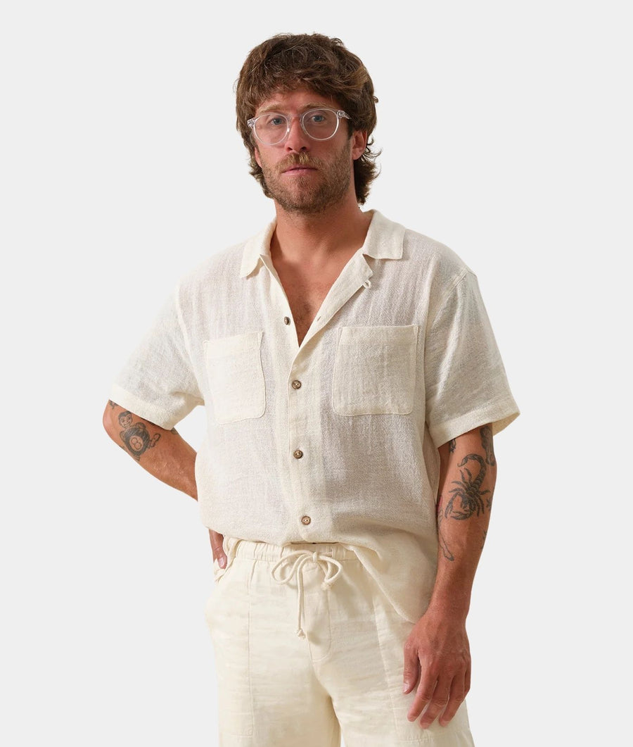 A man wearing a light cream linen short-sleeve button-up shirt and matching drawstring shorts, photographed against a clean light gray studio background.