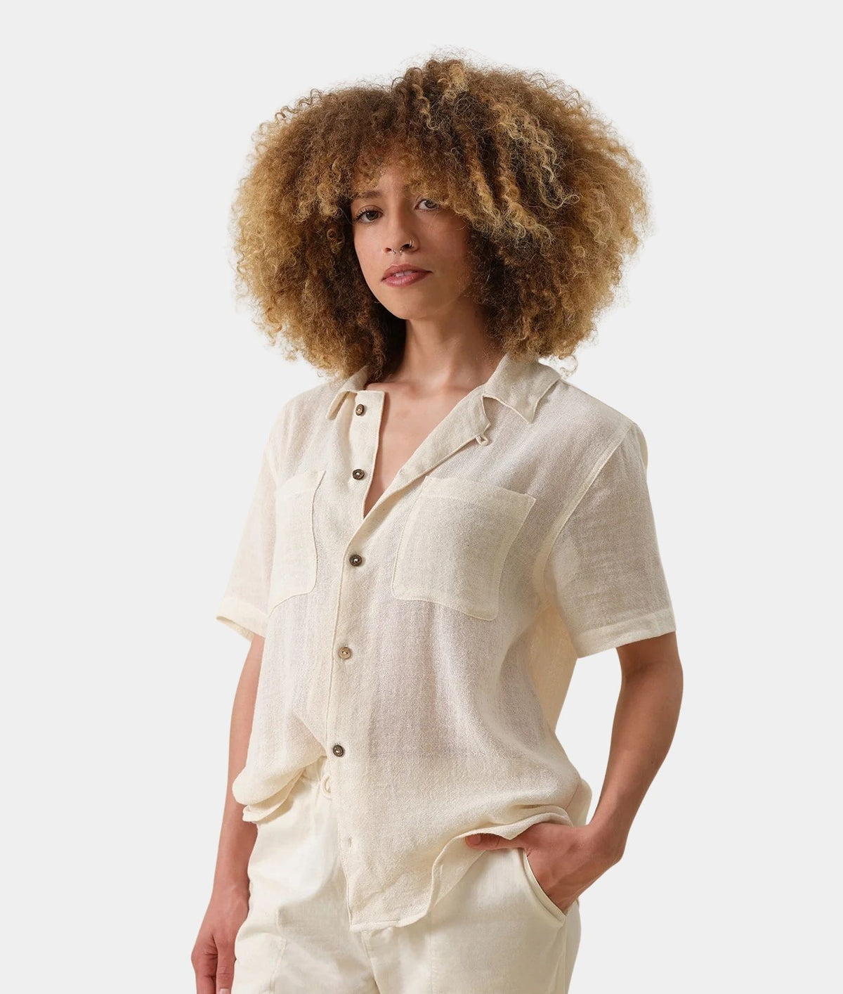 A model with voluminous curly hair wearing a cream-colored short-sleeve linen shirt and matching trousers, photographed against a solid light gray background.