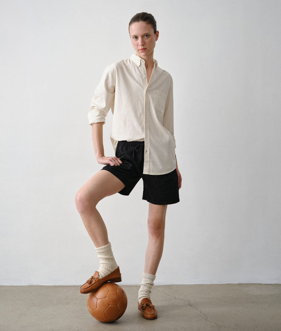 A model stands in a studio wearing a cream long-sleeve linen shirt, black shorts, cream socks, and brown leather loafers, with one foot resting on a brown soccer ball. The background is a clean white wall and concrete floor.