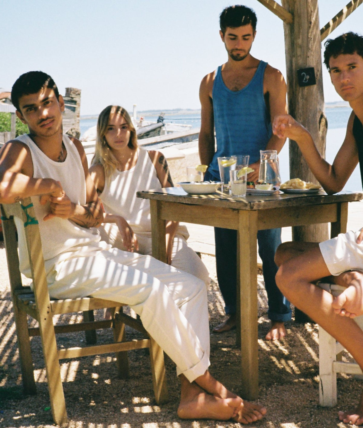 Four people sitting and standing around a wooden table at an outdoor beachside cafe, wearing light-colored tank tops and shorts. The scene features natural sunlight and shadows, with a wooden post and ocean in the background.