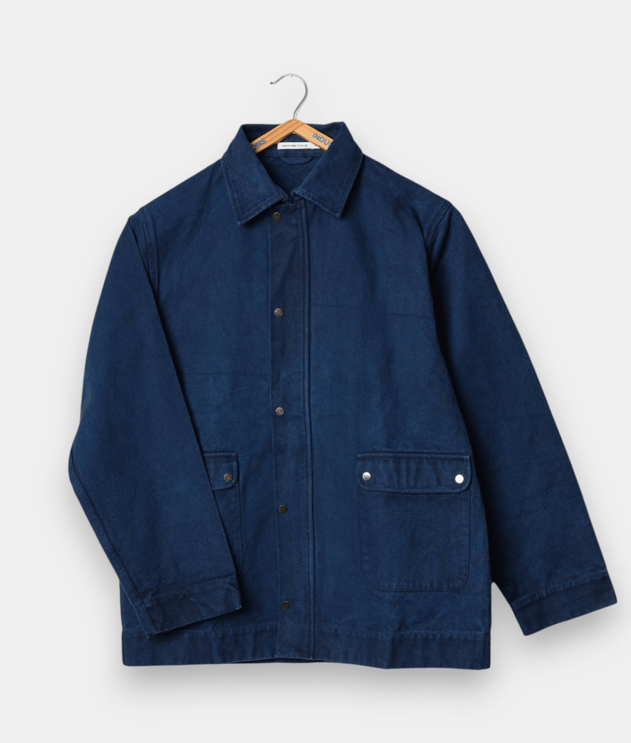 Navy blue denim chore coat hanging on a wooden hanger against a white background - Industry of All Nations