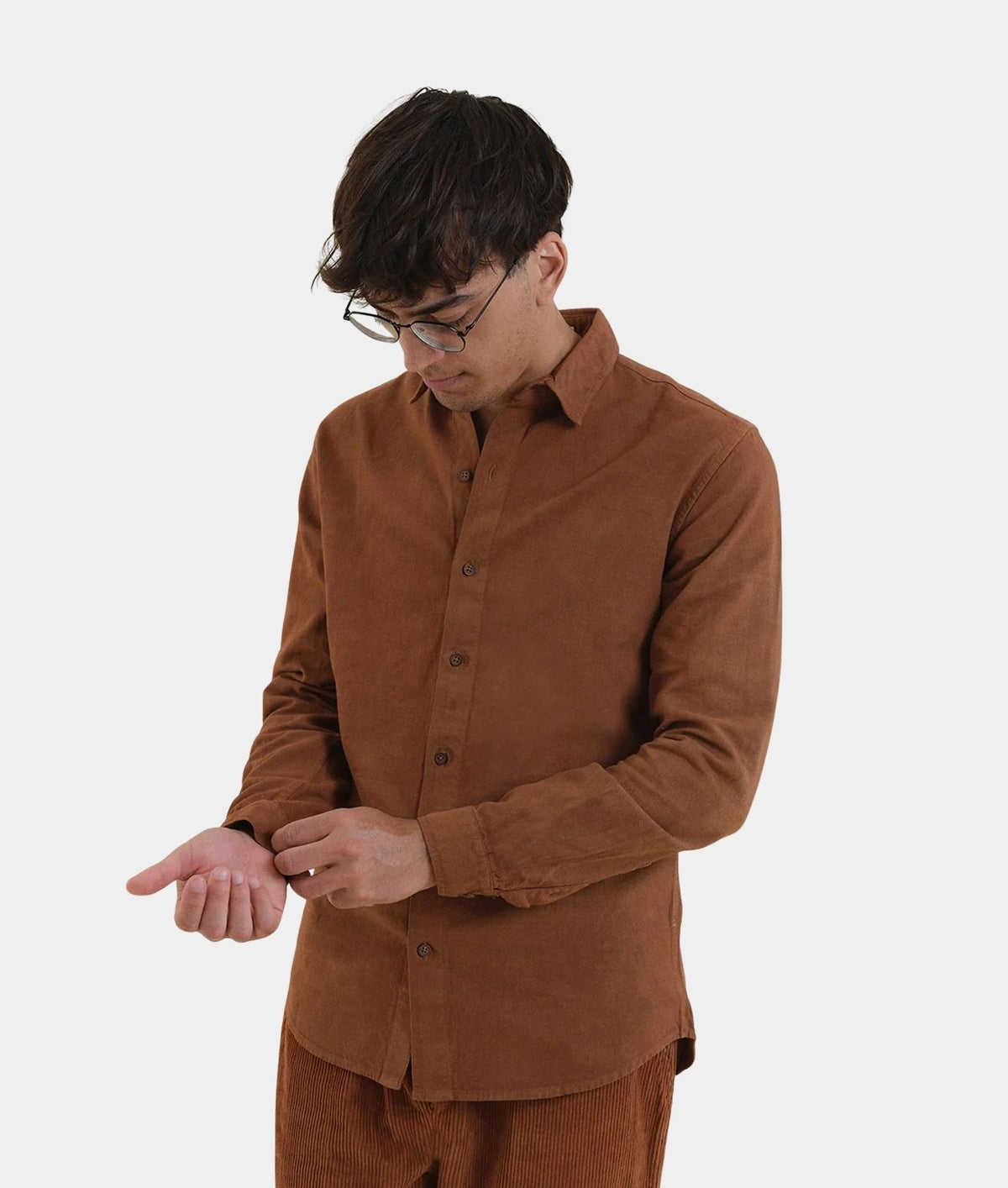 A man wearing a brown corduroy button-down shirt and matching brown corduroy trousers, looking down while adjusting his shirt cuff against a plain light gray background.