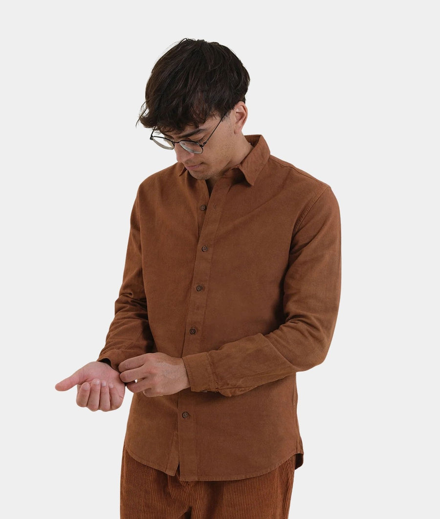 A man wearing a brown corduroy button-down shirt and matching brown corduroy trousers, looking down while adjusting his shirt cuff against a plain light gray background.