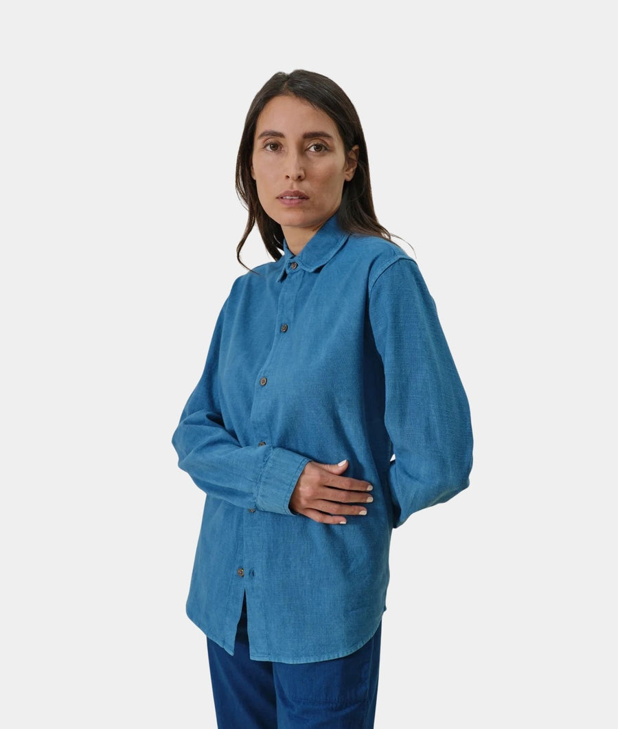 A woman modeling a blue long-sleeve button-up shirt with brown buttons, paired with dark blue pants, against a light gray studio background.