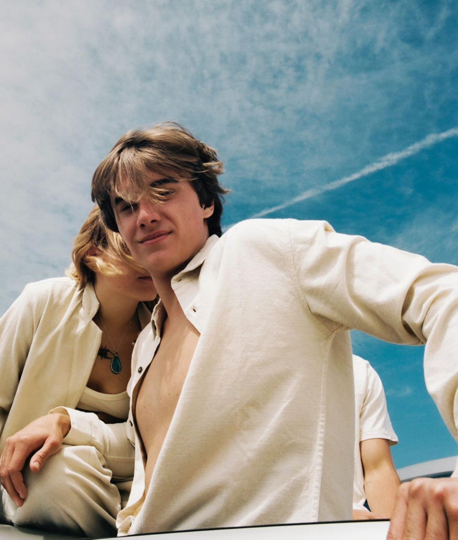 A man wearing an IOAN Pondi Shirt in Undyed leans forward against a blue sky, with a woman resting her head on his shoulder. They wear matching light cream linen clothing.