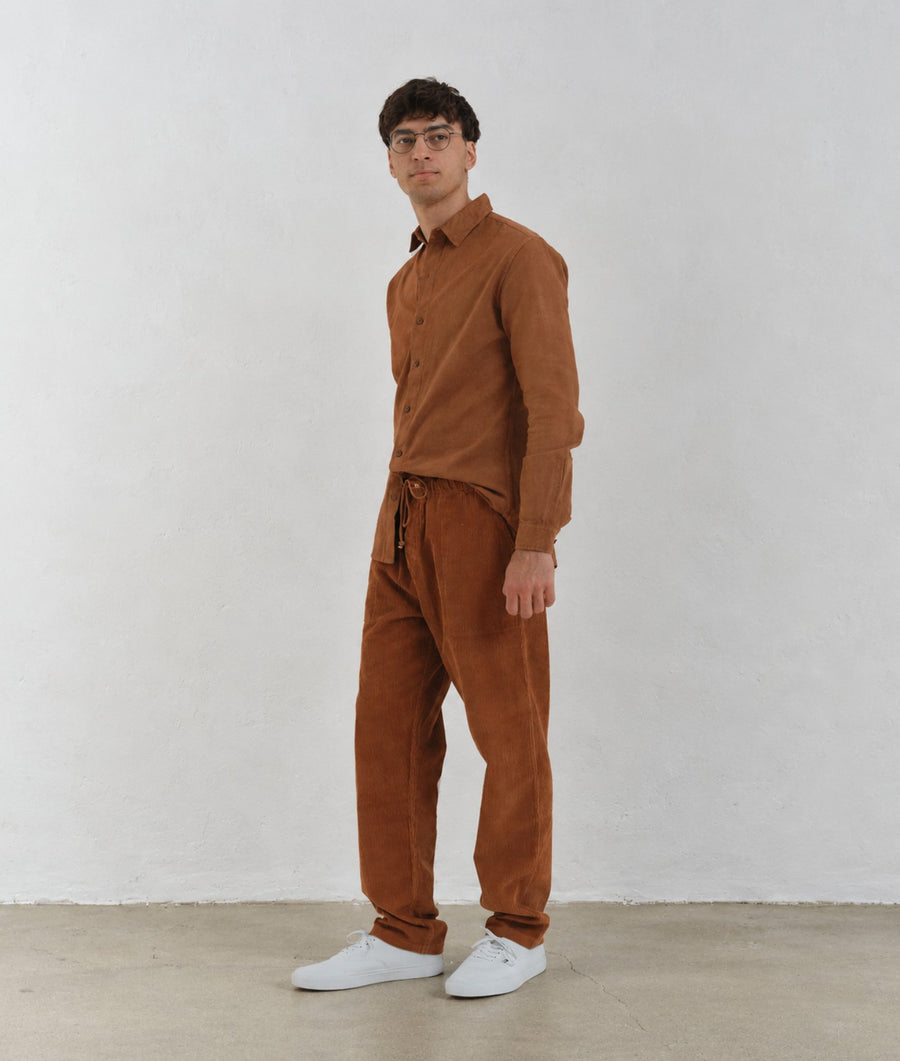 A man modeling the IOAN Ponya Corduroy Pants in Acacia, paired with an IOAN Madras Shirt, against a plain light gray background.