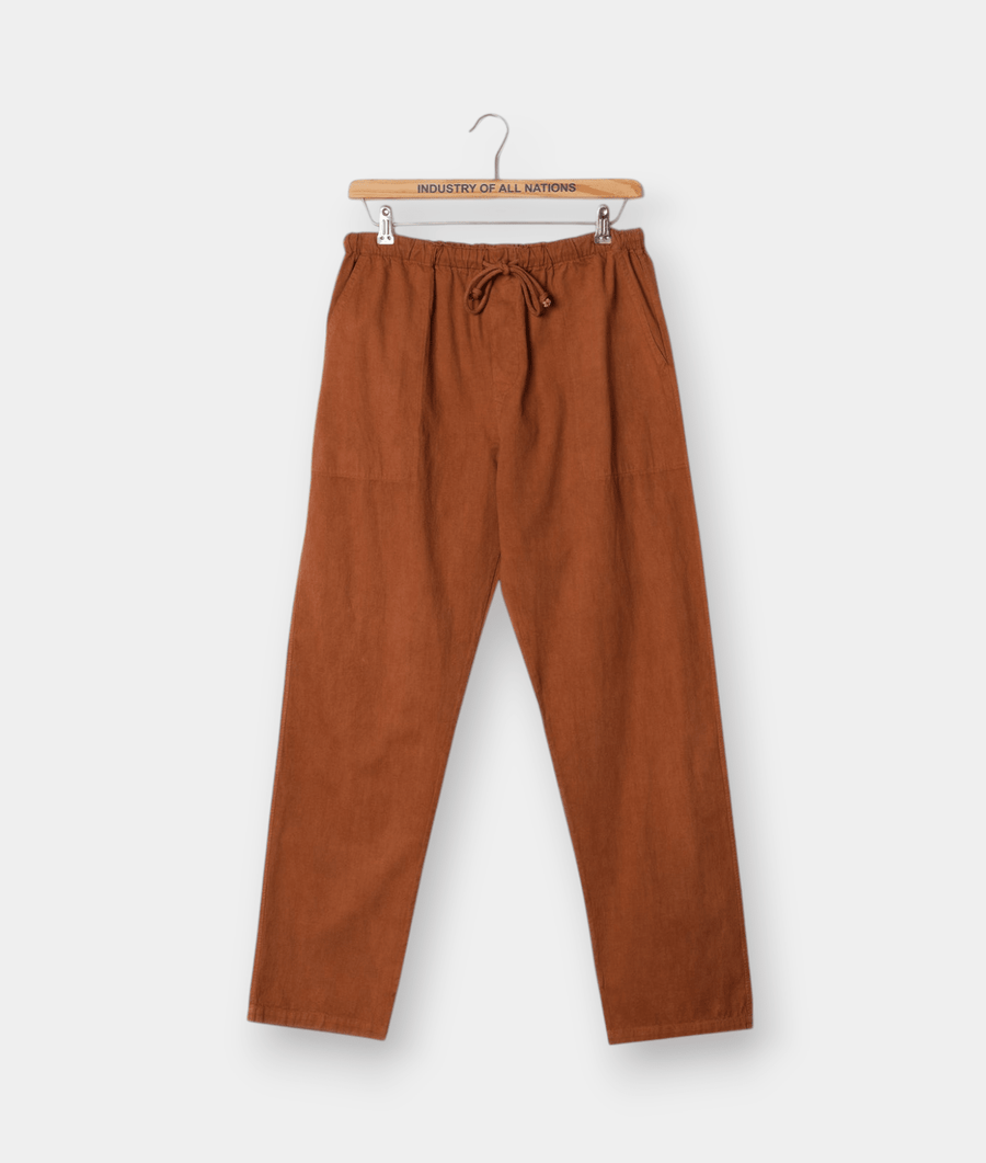 A pair of rust-colored, drawstring linen pants displayed on a wooden hanger with text "Industry of All Nations" - Industry of All Nations