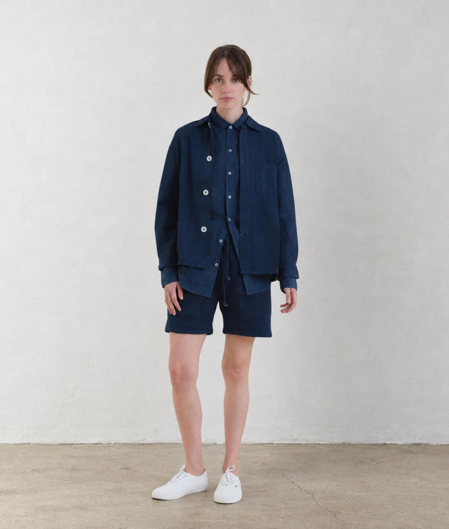 A woman modeling the IOAN Ponya Jacket in Indigo 12, paired with an IOAN Madras Shirt and shorts, against a plain light gray background.