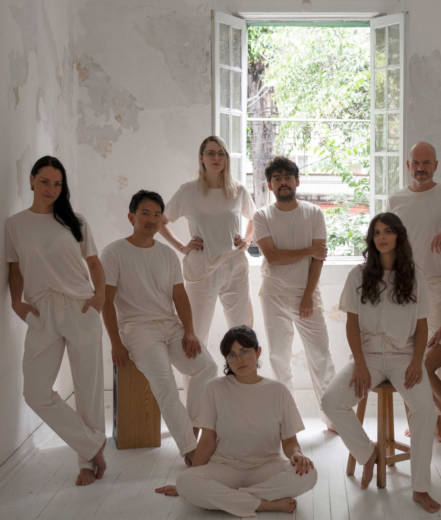 Seven people wearing matching IOAN Clean Crewneck T-shirts and Ponya Drawstring Pants in Undyed pose together in a bright, minimalist room with a large open window overlooking greenery.