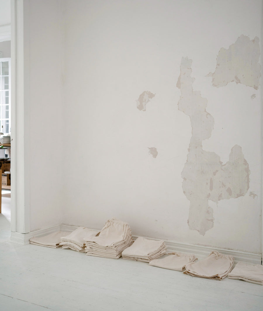A minimalist interior scene showing several folded light beige fabric items arranged along a white floor against a white wall with peeling plaster patches. No person or animal is present, and the image reads as a styled product still life rather than a real-use environment.